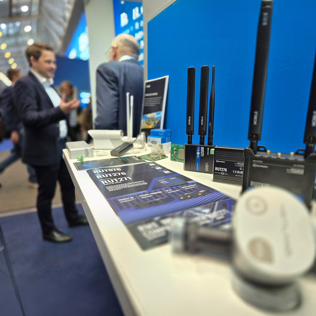 Messestand mit ausgestellten Mobilfunk-Routern und Antennen (z. B. RUT276) auf einem Tisch. Im Hintergrund Besucher im Gespräch.
