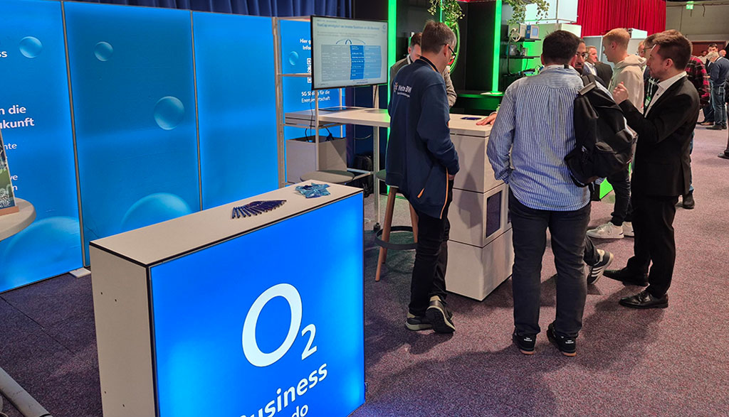 O₂ Business Messestand auf den Metering Days 2025 in Fulda: Mehrere Besucher im Gespräch am Stand mit blauer O₂ Business Branding-Wand. Auf dem Tisch im Vordergrund liegen 5G RedCap-Router zur Demonstration.