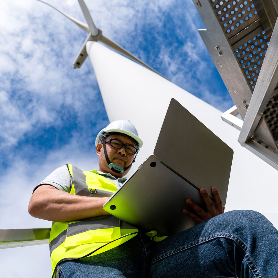 Techniker mit Schutzhelm und Warnweste arbeitet mit Laptop an einem Windradmast