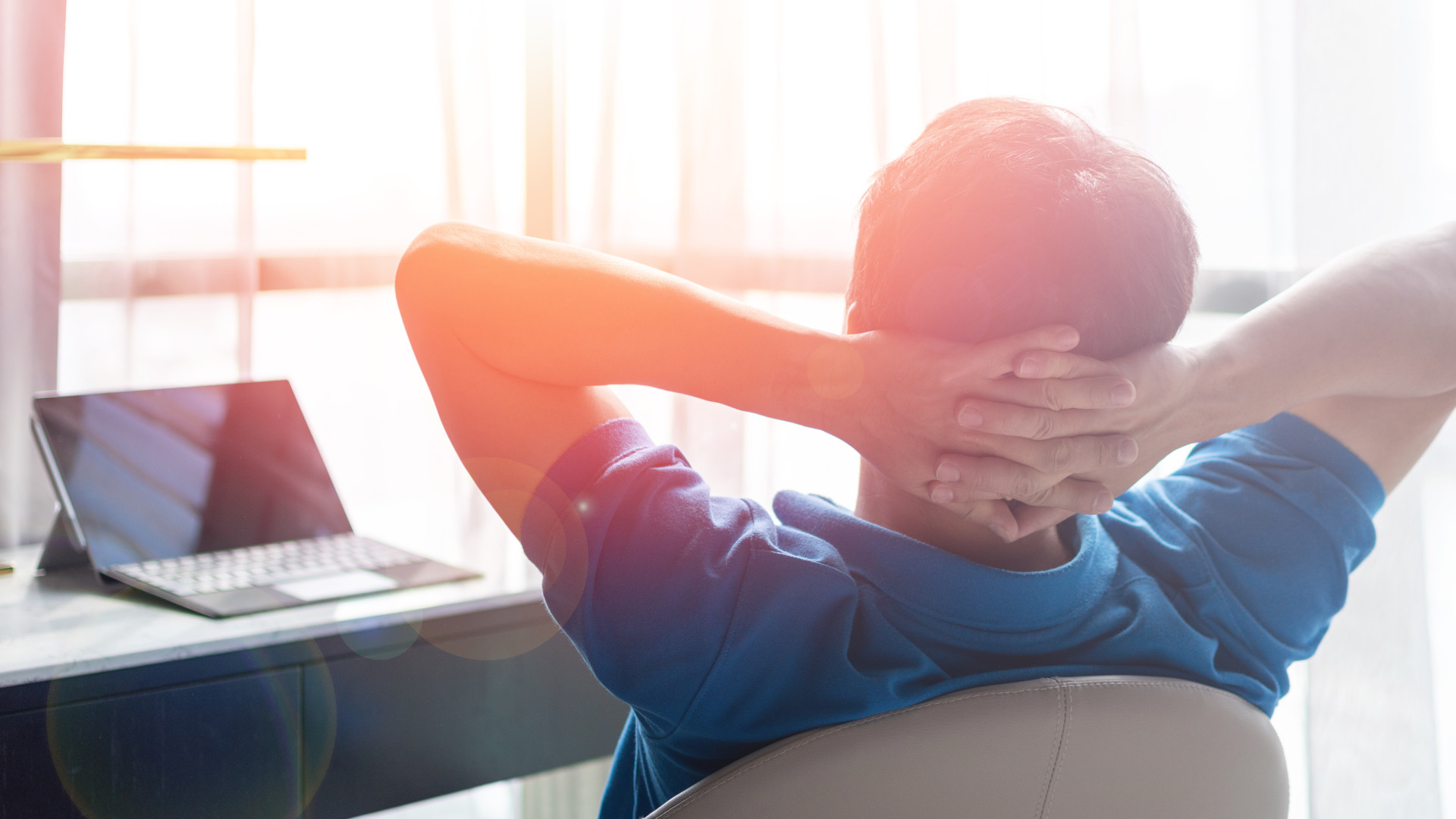 Ein Mann sitzt mit hinter dem Kopf verschrenkten Armen am Schreibtisch und schaut aus dem Fenster.