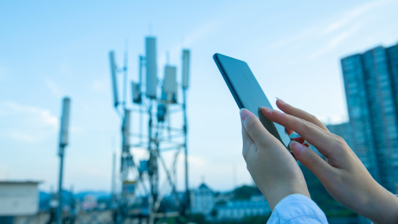 Nahaufnahme menschlicher Hände, die ein Smartphone bedienen. Im Hintergrund ein Mobilfunkmast.