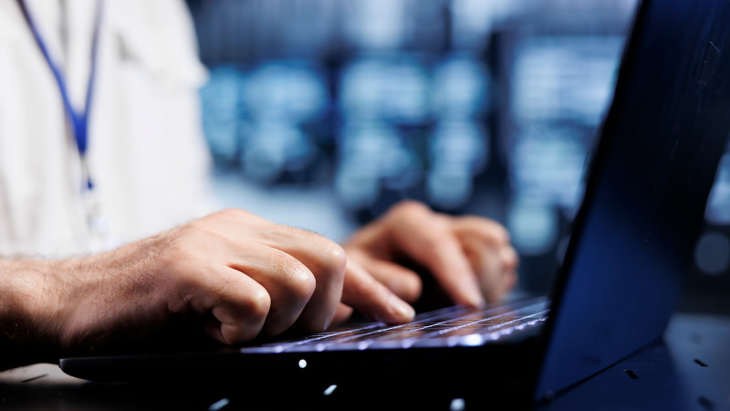 Nahaufnahme von Händen auf einer Laptop-Tastatur. Im Hintergrund ein Serverraum