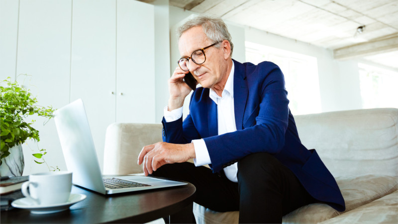 Ein Mann im Anzug arbeitet auf dem Sofa mit Smartphone und Laptop