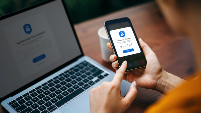 Eine Frau sitzt vor einem Laptop und nutzt Zwei-Faktor-Authentifizierung auf einem Handy