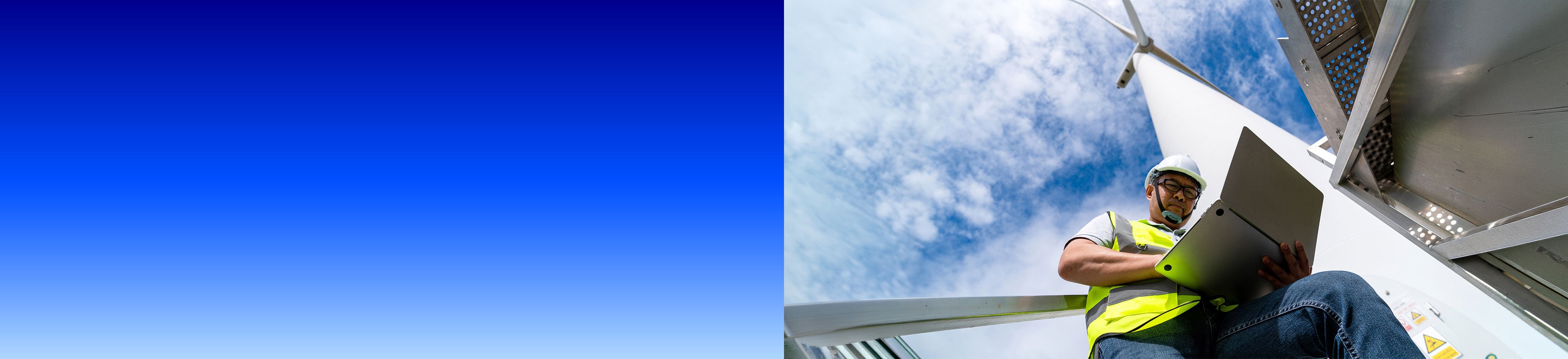 Techniker mit Schutzhelm und Warnweste arbeitet mit einem Laptop an einer Windkraftanlage, Froschperspektive mit blauem Himmel und Wolken, links blauer Farbverlauf.