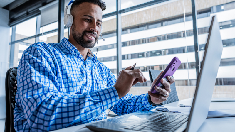 Abbildung: Mann mit Headset sitzt vor einem Computer Monitor und hält ein Smartphone in der Hand