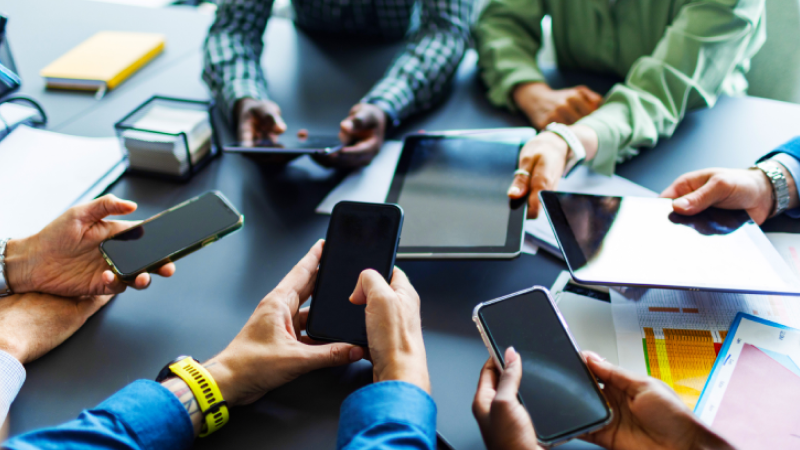 Mehrere Personen sitzen an einem Tisch und halten Smartphones und Tablets, während Unterlagen und Diagramme vor ihnen liegen.