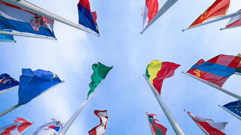Abbildung: Mehrere internationale Flaggen an Fahnenmasten vor blauem Himmel
