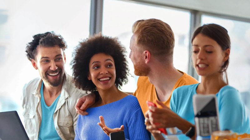 Abbildung: Vier junge Erwachsene sitzen eng beieinander in einem hellen Raum und wirken in ein Gespräch vertieft.  Erste Person links: trägt ein helles Hemd über einem türkisfarbenen T-Shirt, kurze dunkle, leicht gewellte Haare. Zweite Person: blauer Strickpullover, voluminöses, lockiges Haar. Dritte Person: senfgelbes T-Shirt, kurzes blondes Haar. Vierte Person: hellblaues Oberteil, lange glatte Haare, hält ein Smartphone.