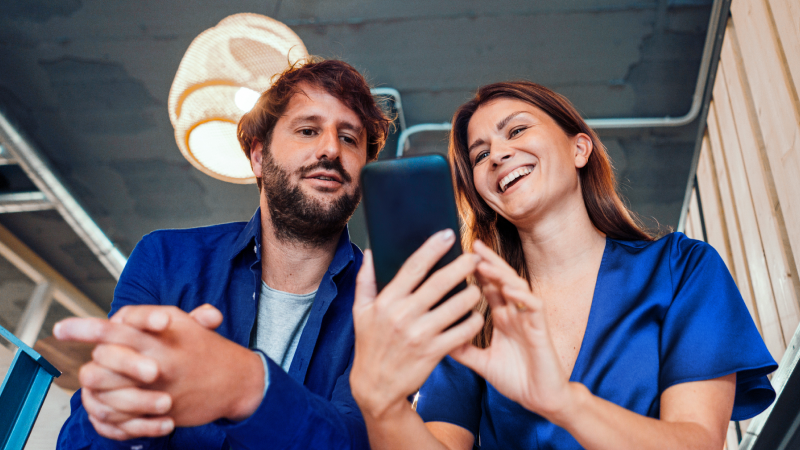 Abbildung: Zwei Personen sitzen nebeneinander in einem modernen Innenraum und schauen gemeinsam auf ein Smartphone, das von der rechten Person gehalten wird. Die linke Person wirkt männlich, trägt ein blaues Hemd über einem hellen T-Shirt und hat kurze, leicht gewellte Haare. Die rechte Person wirkt weiblich, trägt ein kurzärmliges, elegantes Oberteil in kräftigem Blau und hat lange, glatte Haare. Im Hintergrund sind eine Holzverkleidung an der Wand, sichtbare Leitungen an der Decke und eine große, runde Deckenlampe zu erkennen.