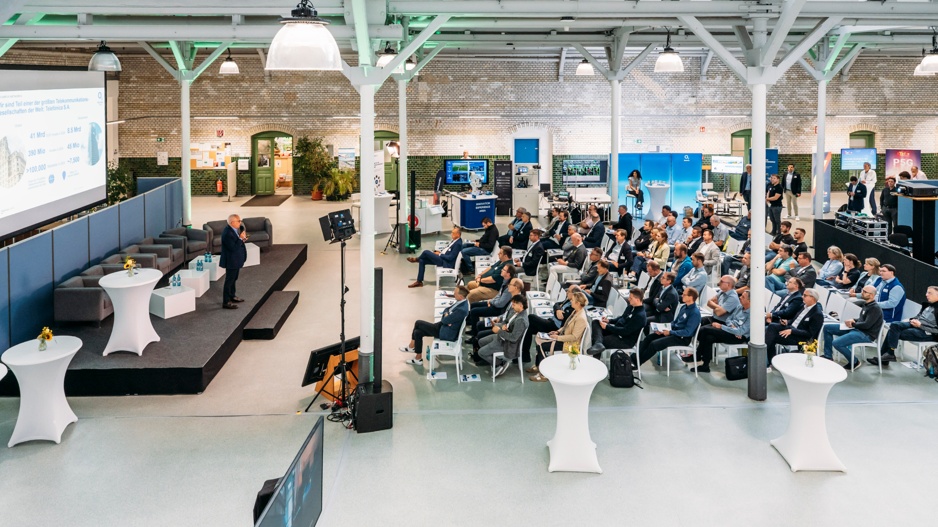Konferenz mit Präsentation auf Bühne, Redner vor Leinwand, Publikum sitzt in Reihen, Backsteinwände und Infostände im Hintergrund.